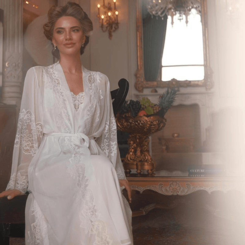 Woman in a white lace robe standing in an elegant room with chandelier and decorative elements.