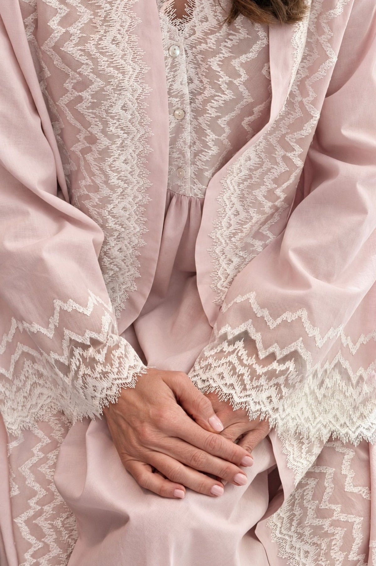 A close-up of wearing a powder yasmine robe with intricate white lace trim along the edges and sleeves, with their hands folded in their lap.