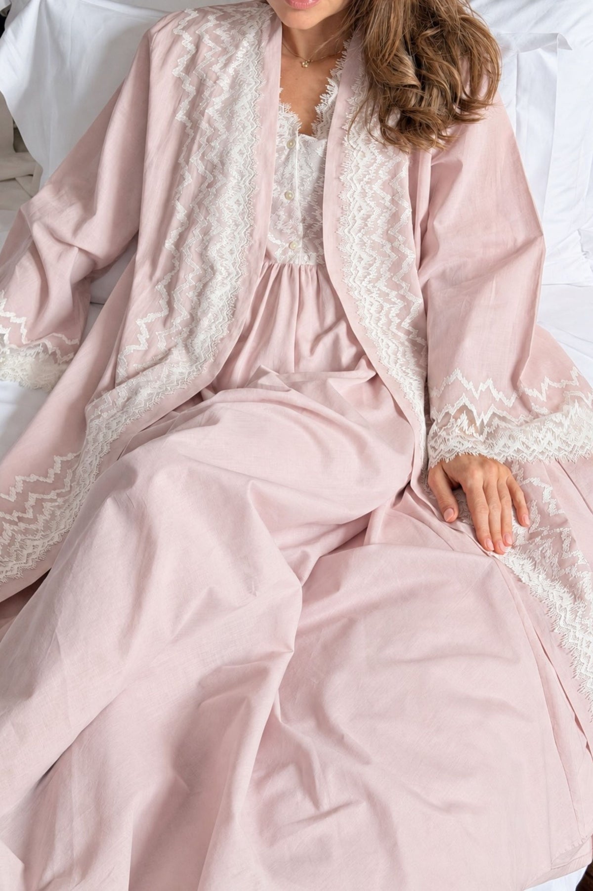 A woman wearing a soft blush pink robe with white lace trim over a matching pink and white nightgown, seated on a white bed.