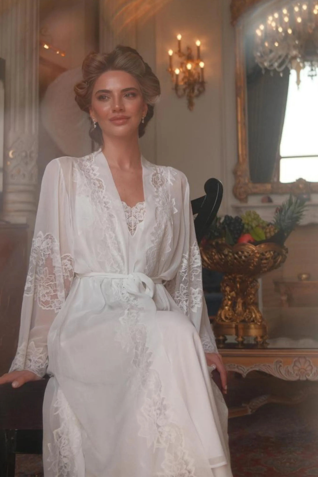 Woman in a white lace robe standing in an elegant room with chandeliers and decorative elements.