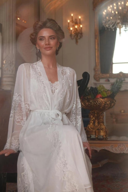 Woman in a white lace robe standing in an elegant room with chandeliers and decorative elements.