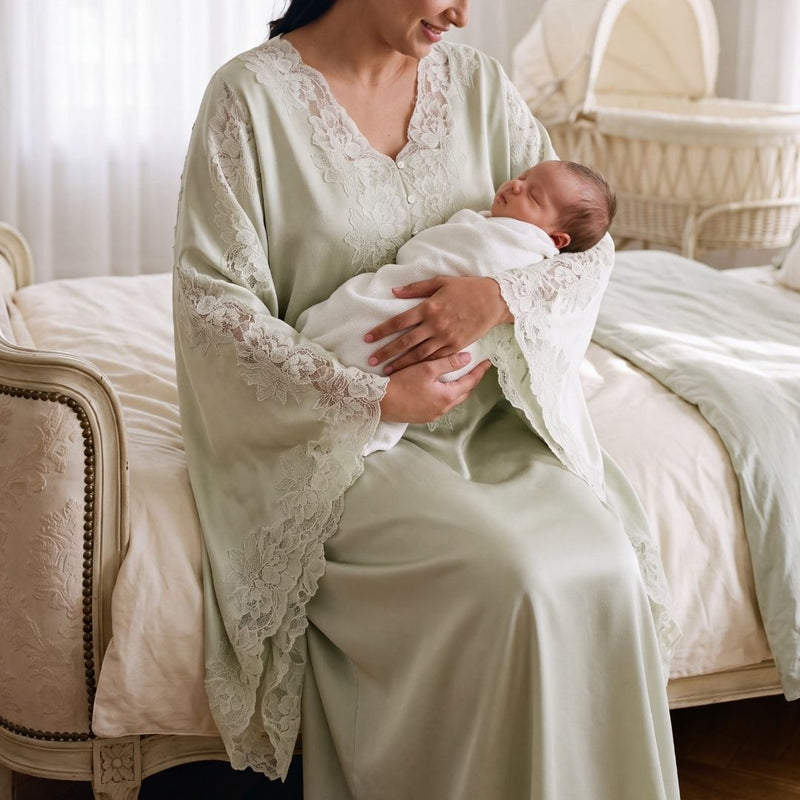 A woman in a sage green satin maternity edit dress with white lace trim holds a sleeping newborn baby wrapped in a white swaddle blanket, sitting on a vintage cream-colored bed.
