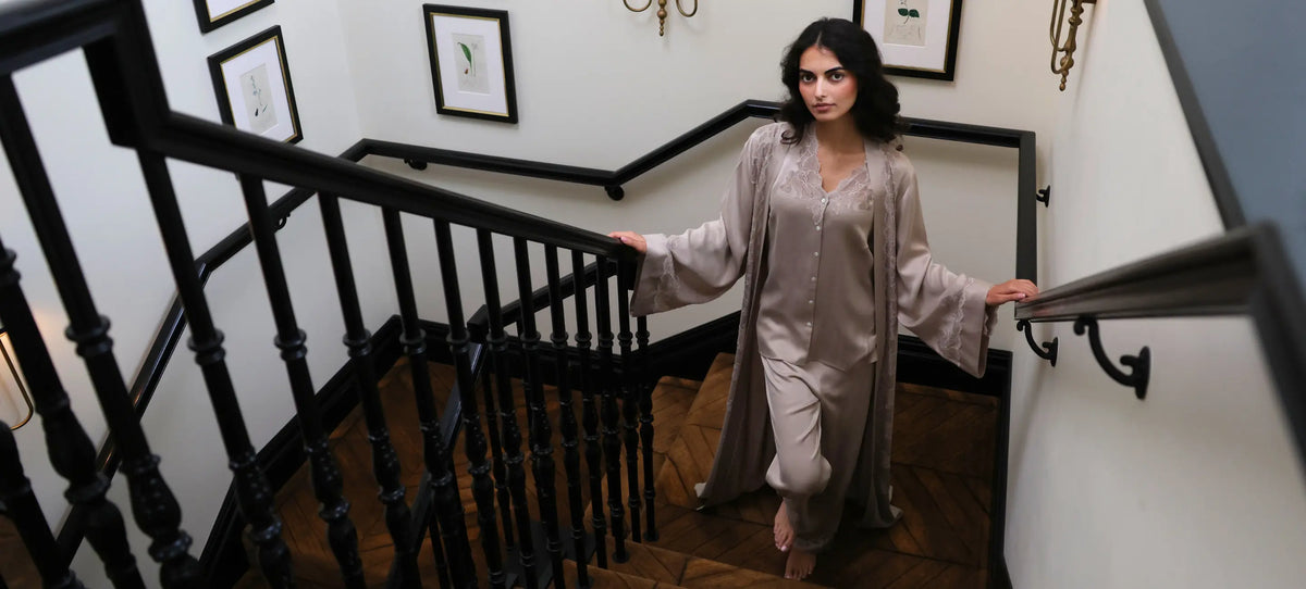 A woman wearing a light taupe satin robe and matching trousers walks down a wooden staircase with a black wooden railing.