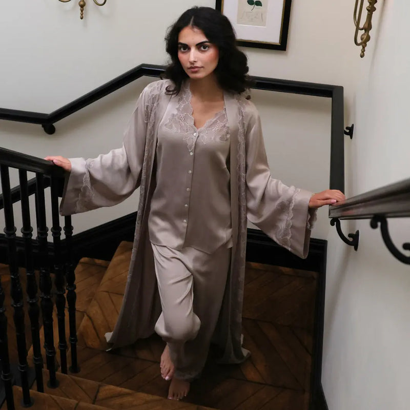A woman wearing a light taupe satin pajama set with a matching embroidered robe stands on a wooden staircase indoors.