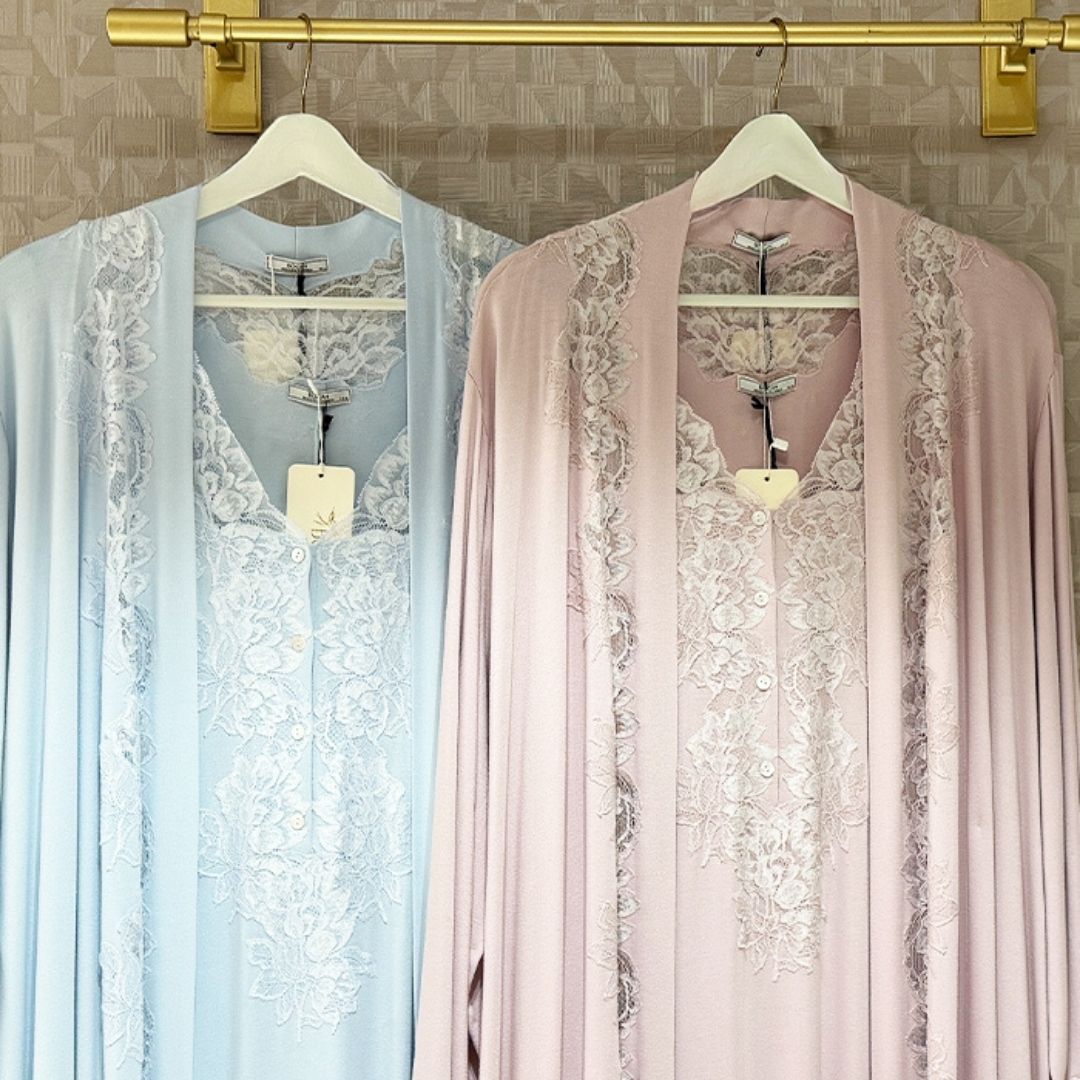 Two lightweight open-front kaftan-style robes hanging on white hangers against a patterned beige wall, one in pale blue and one in pale pink, both with white floral lace panels down the front and along the edges.