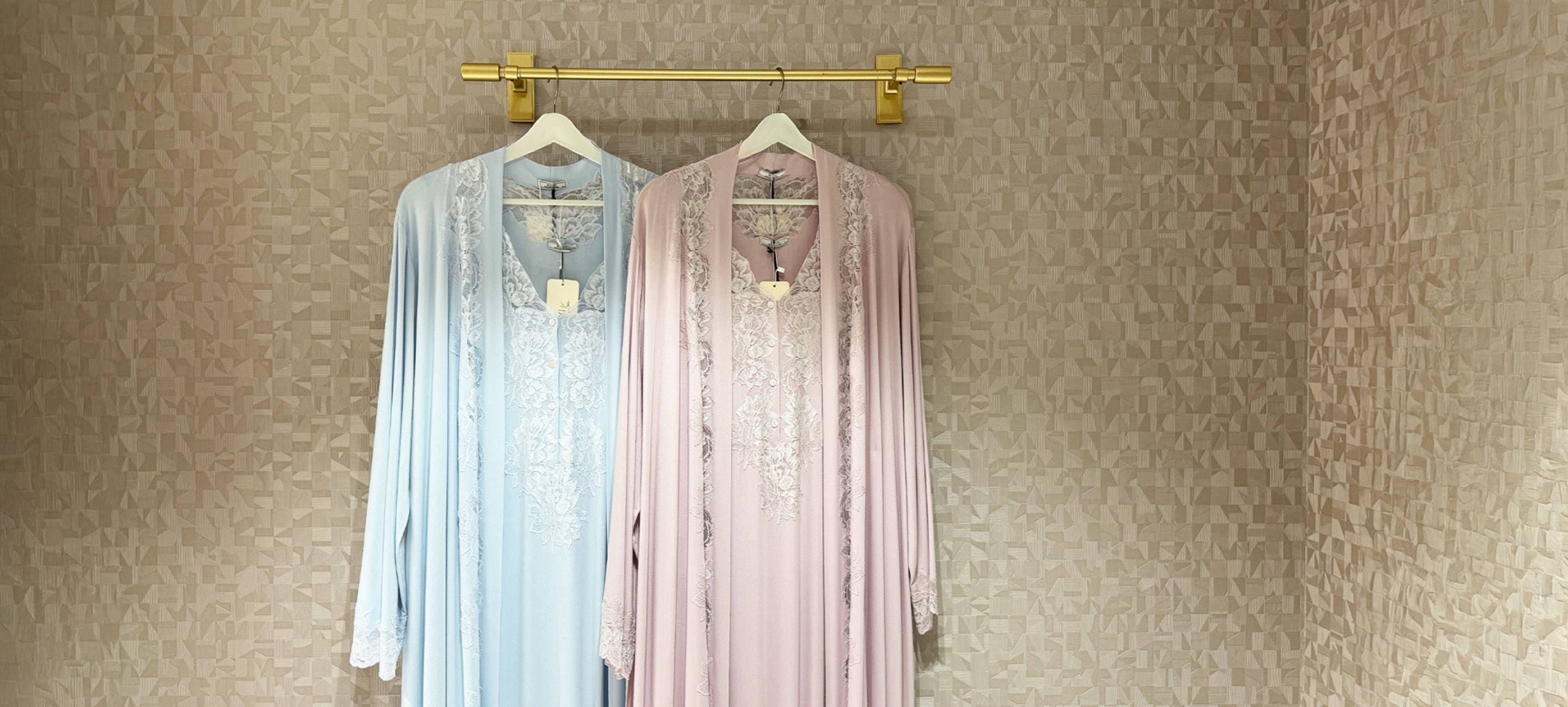 Two long-sleeve embroidered robes hanging on white hangers from a gold wall rod, one in light blue and one in light pink, against a textured beige wallpaper background.