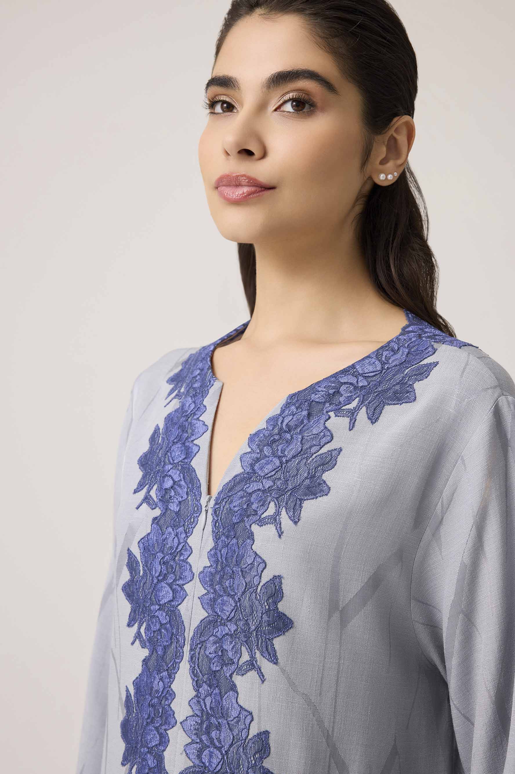 Woman wearing a light gray dress with blue lace details on a plain background