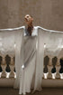 A woman wearing a flowing white satin gown with wide sleeves and a sheer overlay, standing on a stone balcony with her arms outstretched against a warm beige wall.