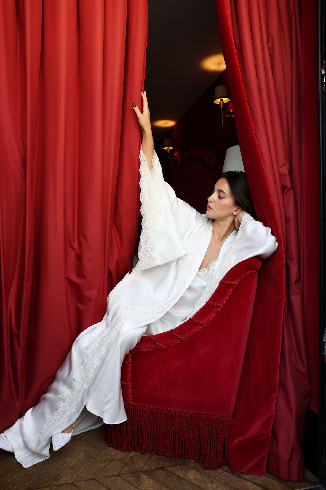 A woman reclining on a red velvet chaise lounge framed by dramatic red curtains, wearing flowing sera white robe.