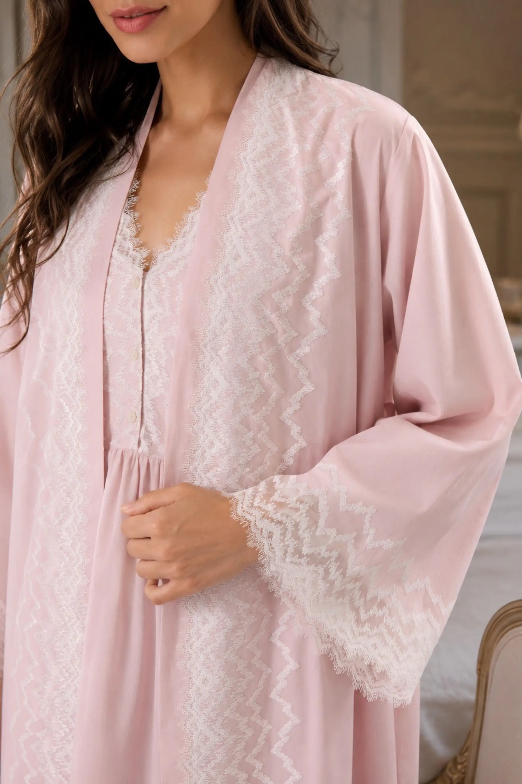 A close-up of a woman wearing a powder robe with white lace trim over a matching powder and white patterned nightgown in an indoor setting.