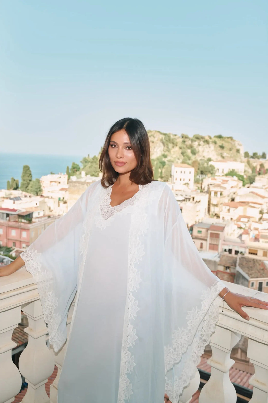 A woman wearing a flowing white lace-trimmed robe stands on a balcony overlooking a coastal town and sea, with her arms resting on the railing.