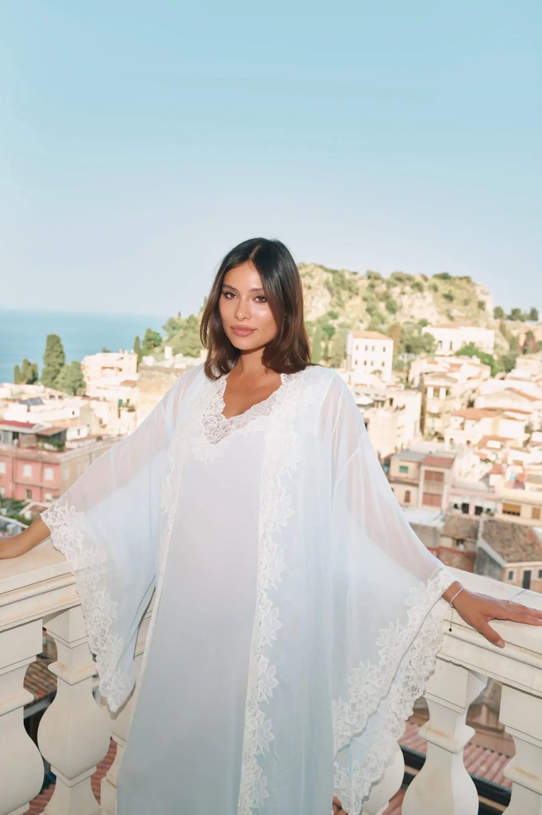 A woman wearing a flowing white lace-trimmed robe stands on a balcony overlooking a coastal town and sea, with her arms resting on the railing.