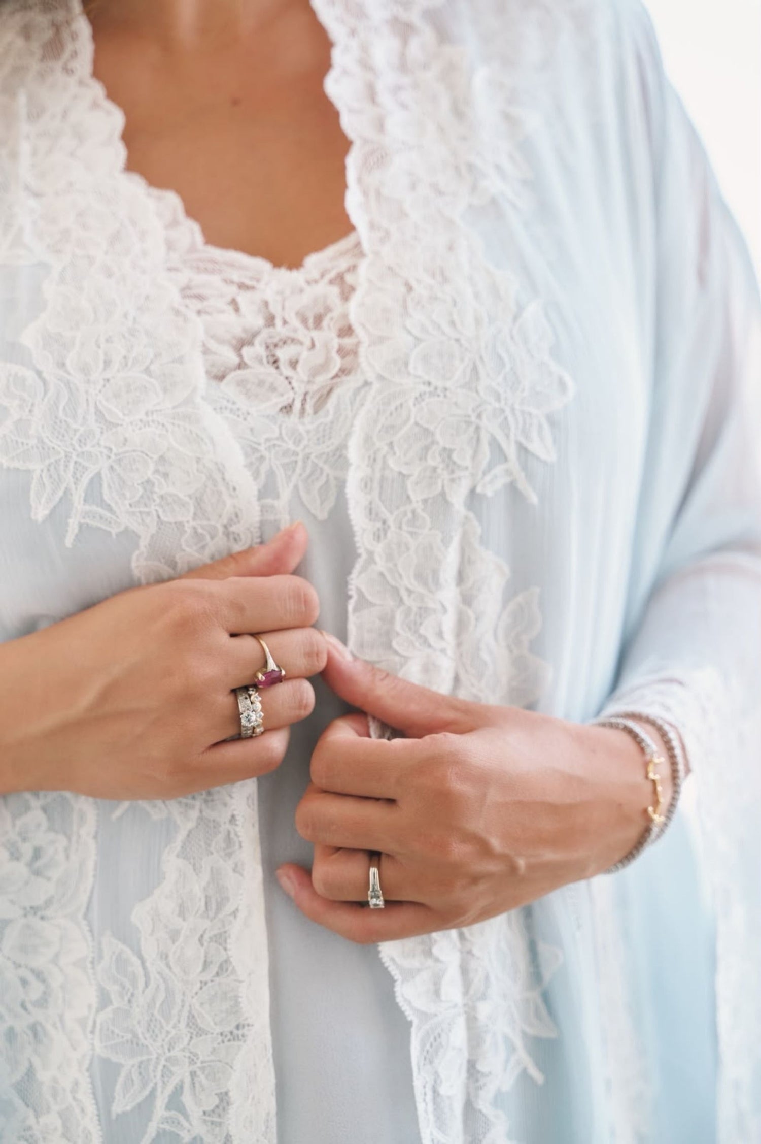 A close-up of a person wearing a light blue sheer robe with white floral lace trim over a matching white lace top, with hands buttoning the robe.