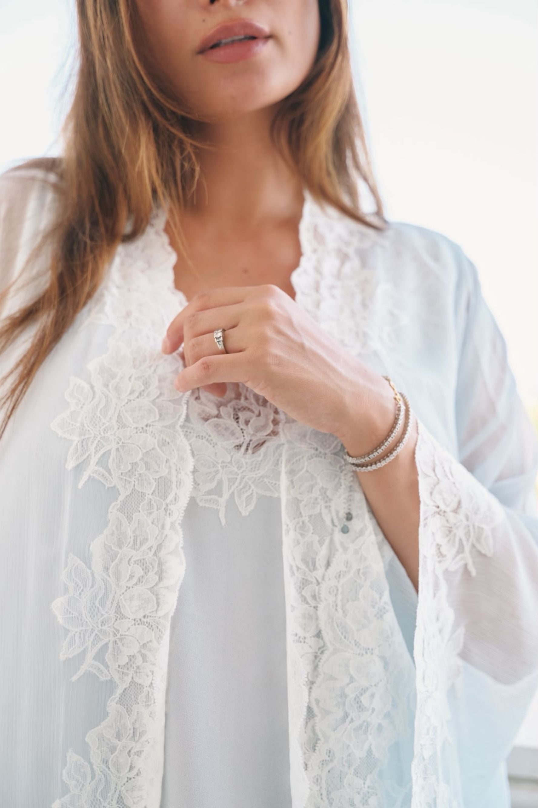 A close-up of a woman wearing a white sheer lace cardigan over a white top, with delicate pearl bracelets on one wrist and a ring on her finger.