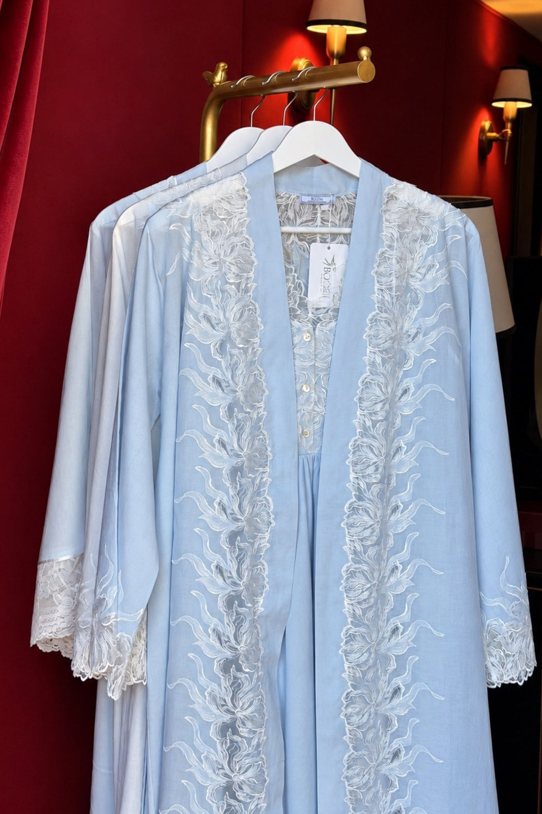 A light blue open-front robe with intricate white floral lace embroidery down the front and on the cuffs, hanging on a white hanger against a dark red curtain background.
