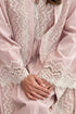 A close-up of wearing a powder yasmine robe with intricate white lace trim along the edges and sleeves, with their hands folded in their lap.